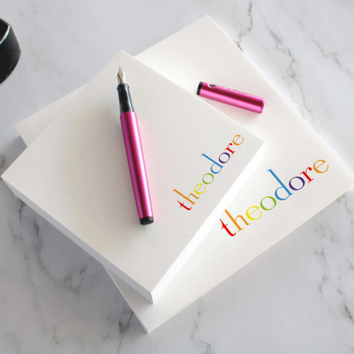 Rainbow square notepads with a rainbow-colored name near the lower corner, beside a pink pen.