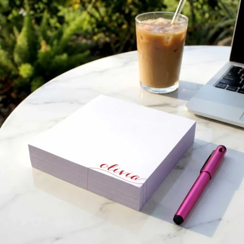 Holla Swash square notepad with a pink script name, on an outdoor table beside iced coffee.