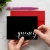 Hands hold a Holla Swash folded notecard with white Quincy script on a black front, with a red envelope and pink pen behind it.