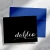 Holla Swash folded notecard with white Dahlia script on a black front, shown in front of a royal-blue envelope on a marble desk.