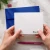 Hands hold a Holla Swash flat notecard with rounded corners and a black swash name, with a royal-blue envelope, pink pen and binder clip on the desk.