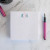 Traditional Script square notepad with pink and teal monogram, shown beside a pink pen on a marble desktop.