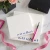 Two Holla Swash square notepads personalized in blue script, arranged with a pink fountain pen, ribbon, flowers and a black box.