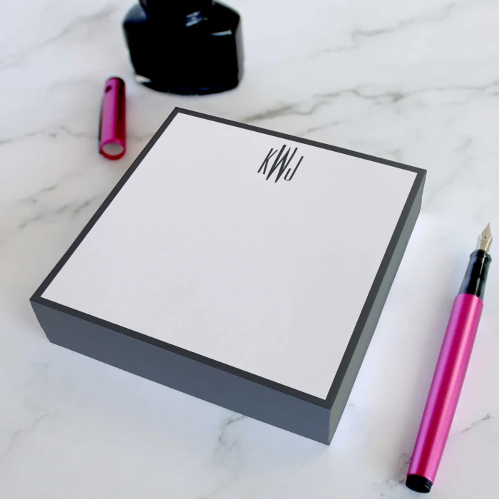 Gray-bordered personalized square notepad with black monogram, pink pen, and ink bottle