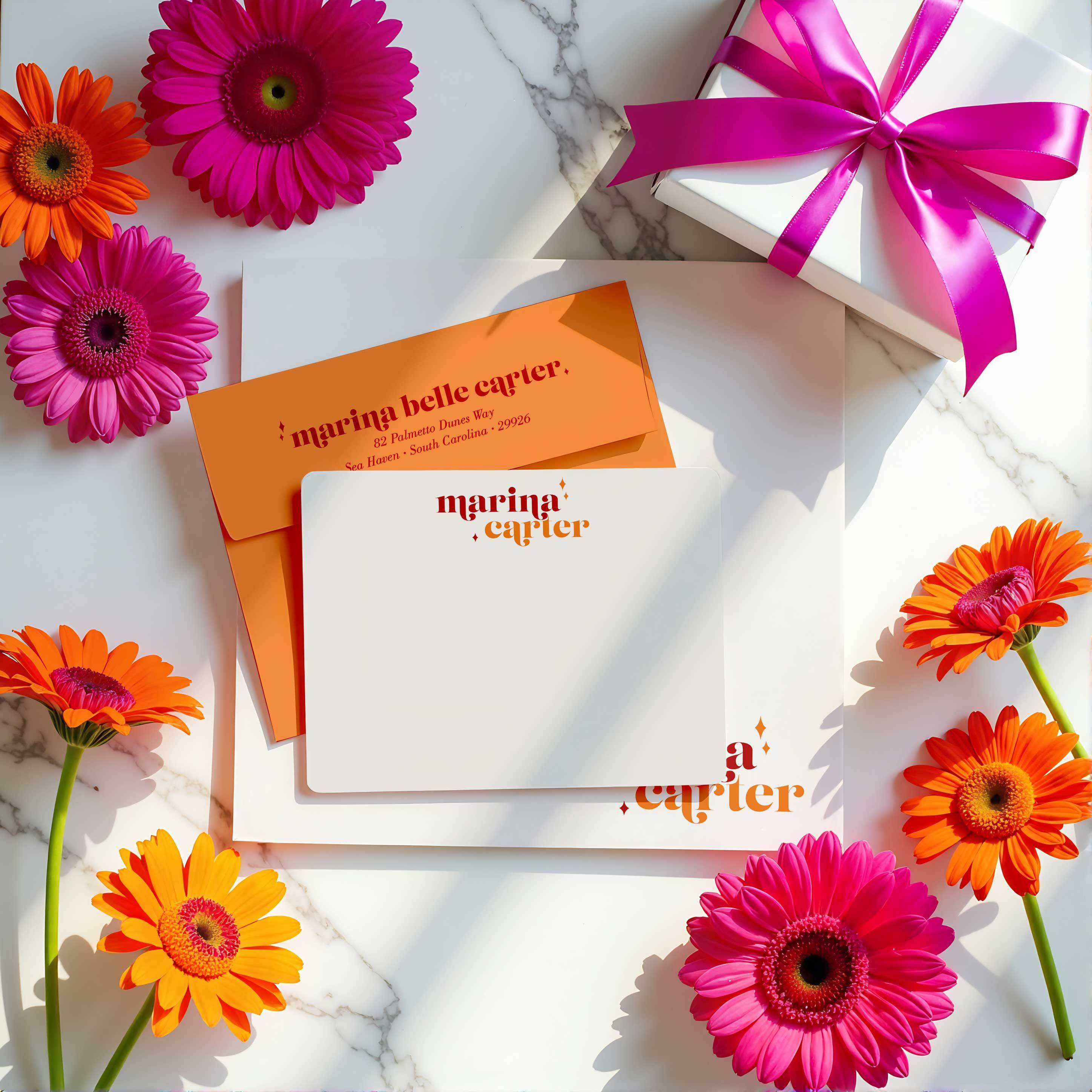 Personalized flat notecards with bold orange and pink lettering, paired with matching return address envelopes and surrounded by bright birthday flowers and a gift box—custom stationery set for her, perfect for birthday gifting.