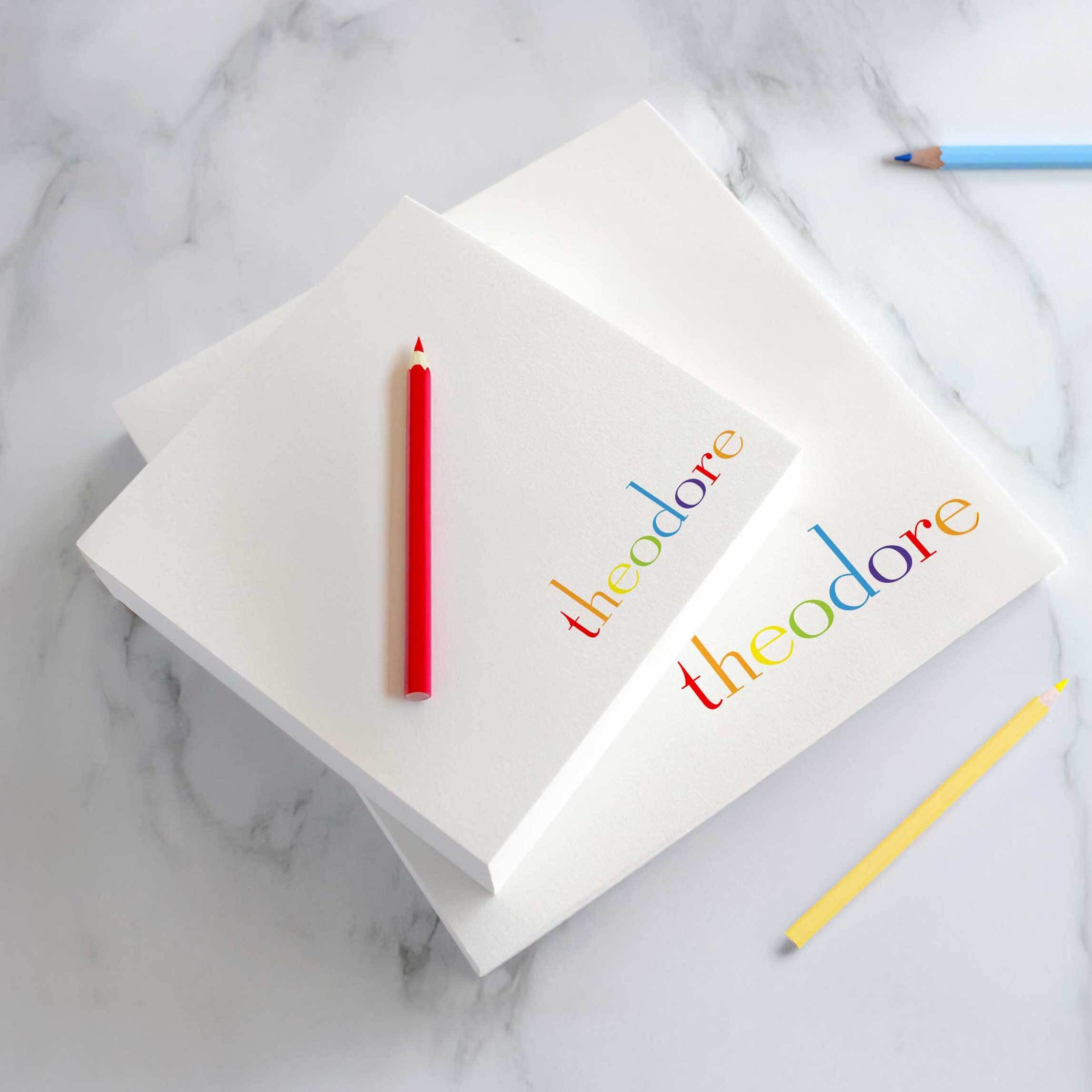 Personalized rainbow name notepad with “Theodore” in multicolor text, custom stationery pad for kids or gifts on marble desk with colored pencils