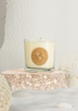 A white soy candle sits atop a rock surrounded by sand and coral.