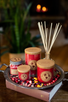 An assortment of red Crushed Cranberry candles and a reed diffuser are arranged on a silver tray with fresh cranberries and orange peel atop a book in a cozy home setting.