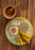 A pale yellow soy candle lays on its side on a floral lunch plate next to juicy slices of fresh mango and a wooden bowl of raw honey.