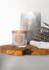 A pale purple soy candle rests on a rustic wooden platter alongside a bunch of dried lavender flowers with a shiny glass candle holder in the background.