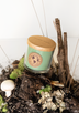 A green soy candle sits atop an earthy display of moss, soil, lichen, mushrooms, and twigs.