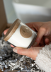 Hands holding a white soy candle with golden label above box with festive packing.