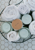 Fresh looking display of pale blue candles snuggled amongst fluffy, white bath towels in a black metal basket on a clean white tile background.