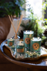 Beautiful arrangement of green soy candles all lit on antique silver serving tray with a reed diffuser and greenery in the background.