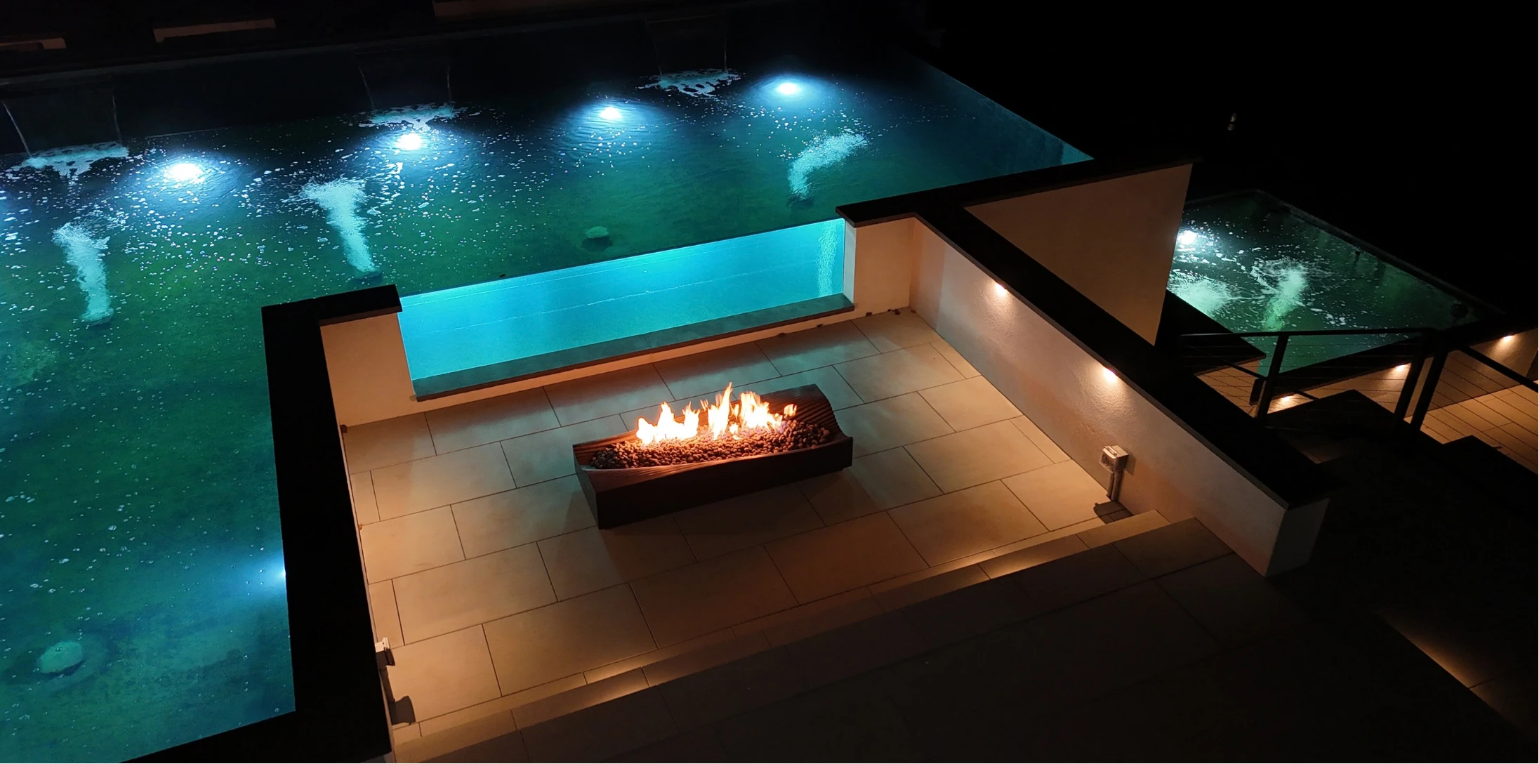 Illuminated glass-walled pond with a fire feature on a modern patio at night