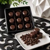 Nine handcrafted Baileys & Coffee bonbons in a premium black gift box with four faceted artisan chocolates and roasted coffee beans displayed on a white serving plate.