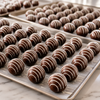 Rows of handcrafted milk chocolate hot cocoa bombs with elegant white chocolate drizzle arranged on sheet pans during production, showcasing Bomb Confections' small-batch craftsmanship and large-order capabilities for weddings, corporate gifts, and event favors.
