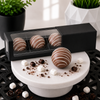 Three handcrafted milk chocolate hot cocoa bombs with elegant white chocolate drizzle displayed in a premium black slider gift box with a clear viewing window, styled with mini marshmallows, chocolate curls, and cocoa powder in Bomb Confections' signature luxury presentation.