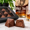 Close-up of handcrafted Buffalo Trace Bourbon Bonbons by Bomb Confections with premium window gift box, subtle Buffalo Trace bourbon bottle, and whiskey glass in the background.