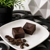 Two handcrafted espresso bonbons with a geometric design, displayed on a white plate with roasted coffee beans. Artisan coffee-infused chocolates by Bomb Confections.