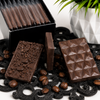 Close-up of Bomb Confections Espresso Mini Chocolate Bars showing the geometric molded front, chopped espresso bean back, and bar thickness alongside roasted coffee beans.