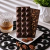Bomb Confections Bourbon Chocolate Bar showing the smooth geometric front alongside the optional chopped pecan back, displayed with subtle Buffalo Trace bourbon accents and pecans.