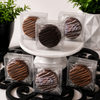 Collection of individually wrapped chocolate covered sandwich cookies in clear gift boxes featuring classic, peanut butter, dark chocolate, and birthday cake varieties displayed on white pedestal stands.