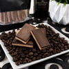 Close-up of Bomb Confections Baileys & Espresso Toffee featuring rich dark chocolate layered over buttery Baileys-flavored toffee, displayed on roasted coffee beans with premium gift pouch packaging softly blurred in the background.