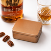 Premium copper gift tin for Bomb Confections Buffalo Trace Bourbon Bonbons, shown closed with elegant gift packaging alongside Buffalo Trace bourbon, a crystal whiskey glass, and pecan halves on a clean white background.