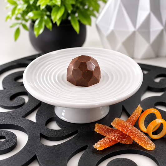 Single handcrafted Bourbon Old Fashioned bonbon by Bomb Confections displayed on a white pedestal with candied orange peel and orange twist accents on a black scroll mat.