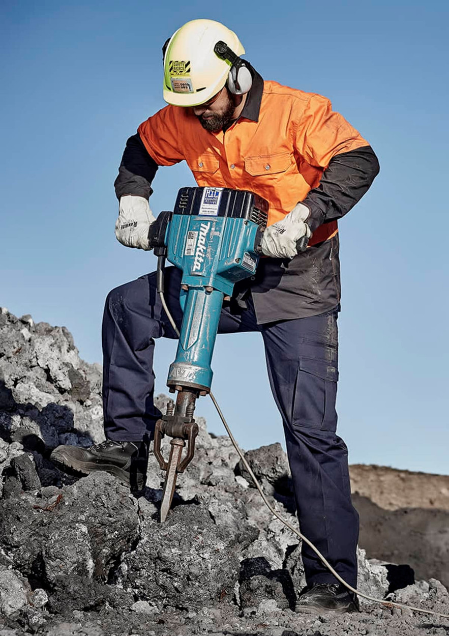 A man in an orange and black jacket and ear protection operates a blue jackhammer on rocky terrain.