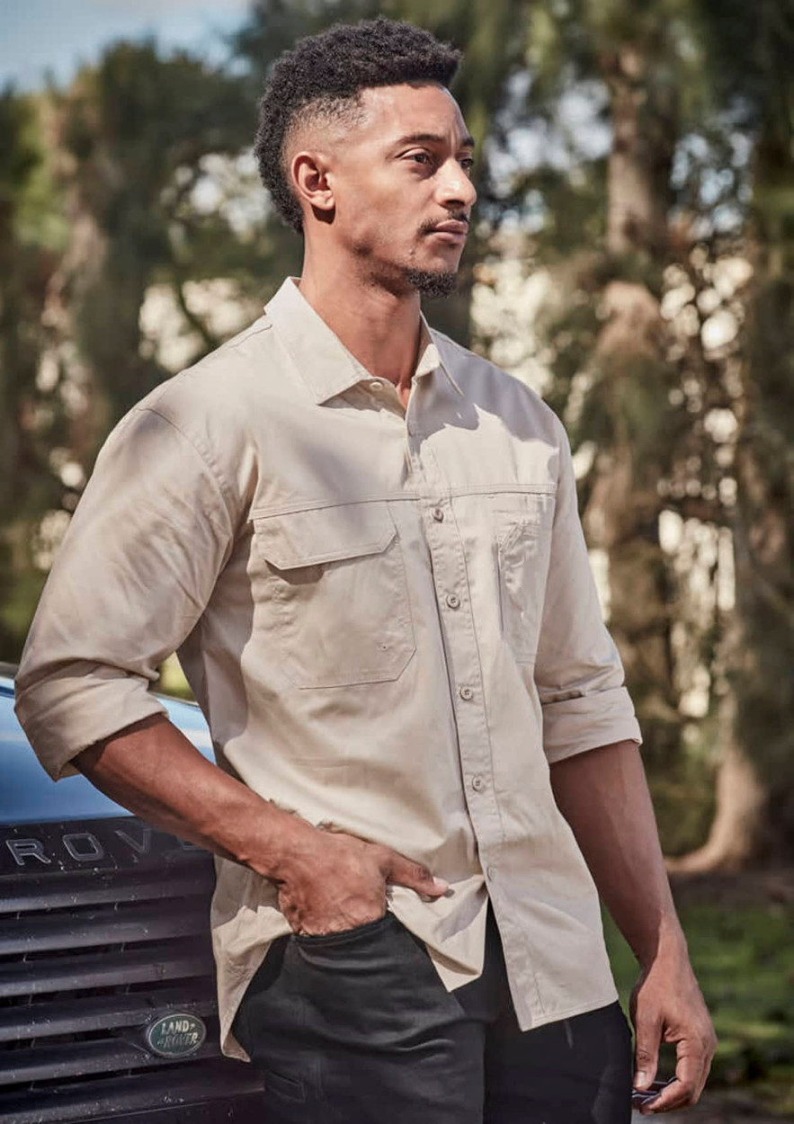 A lightweight long sleeve shirt in beige, featuring a front pocket and a logo. Set outdoors with greenery in the background.