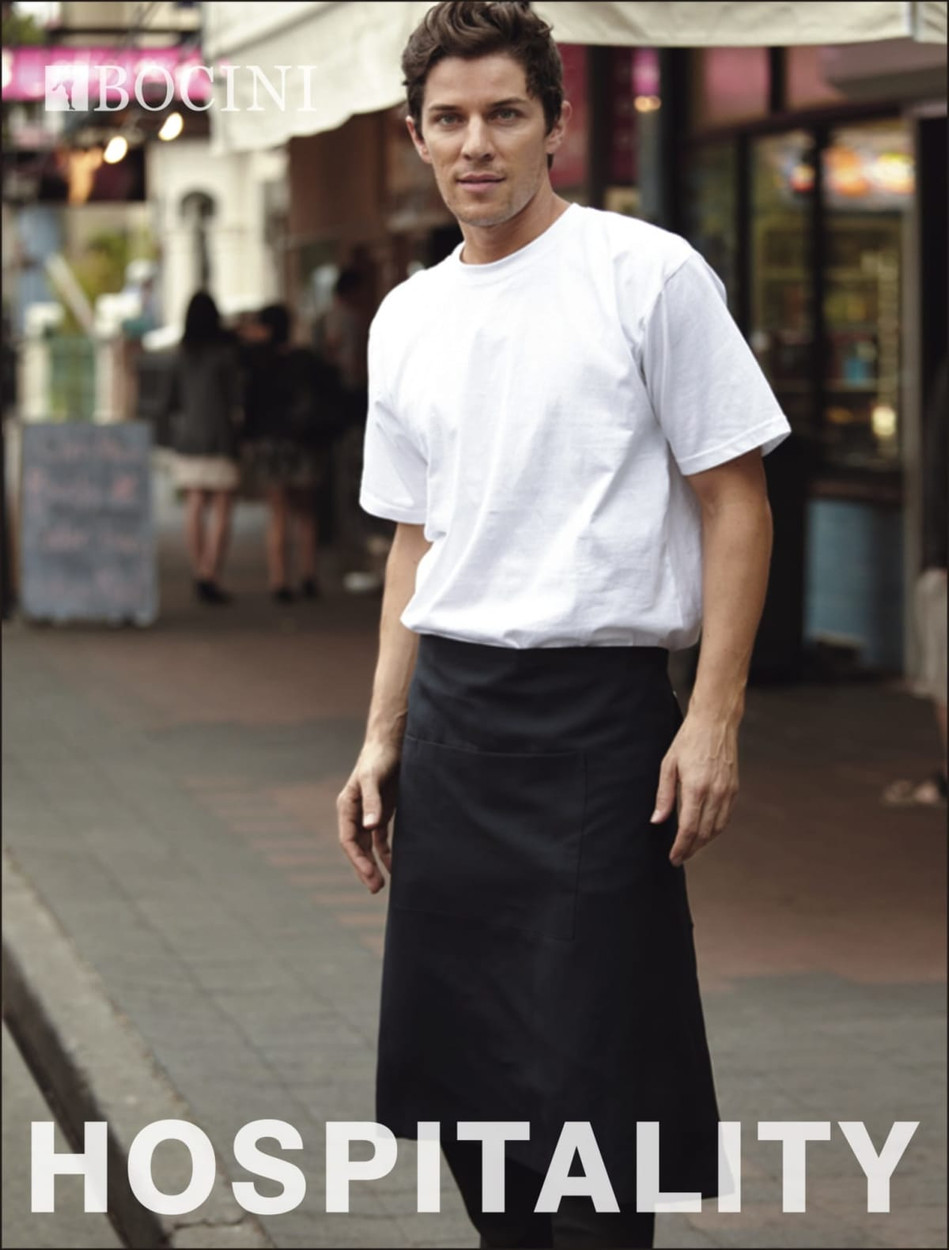 A man wears a black cotton drill three-quarter apron over a white T-shirt, standing in an outdoor setting. The apron features a pocket.