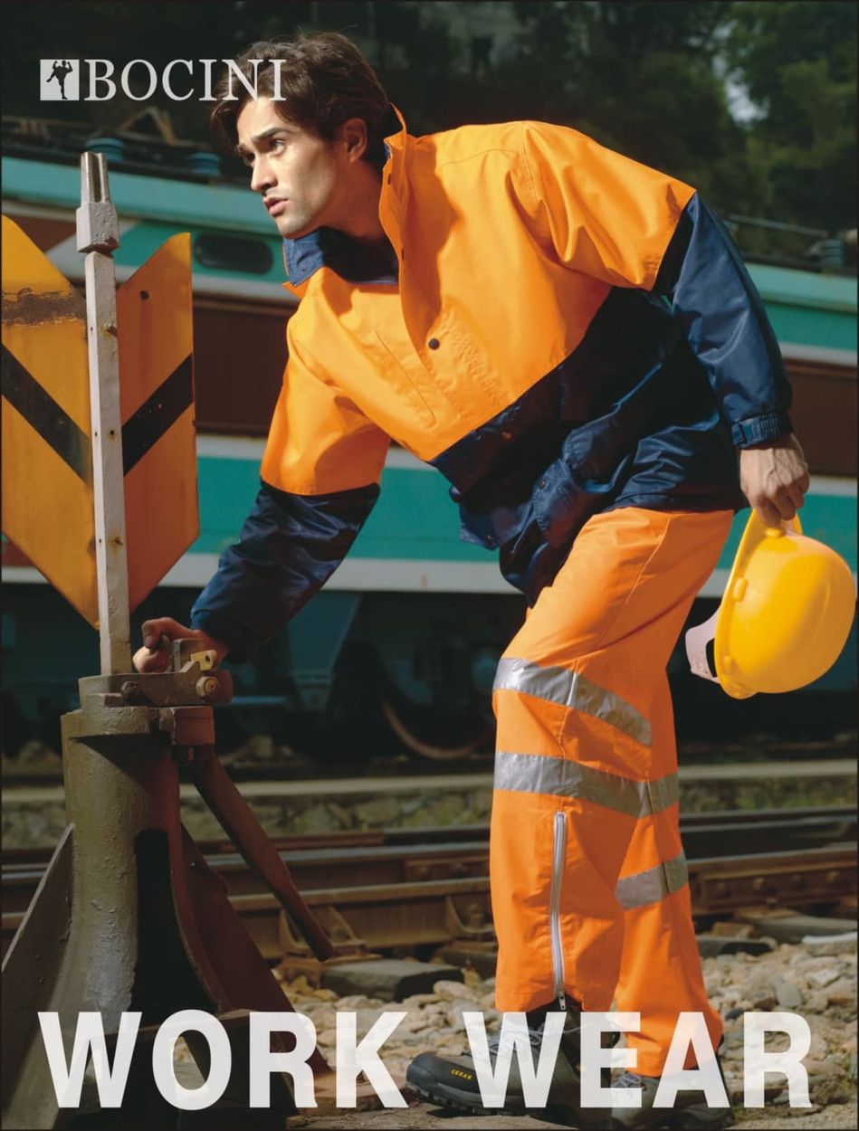 A hi-vis jacket in orange and navy, featuring a reflective design, worn by a model near railway tracks.