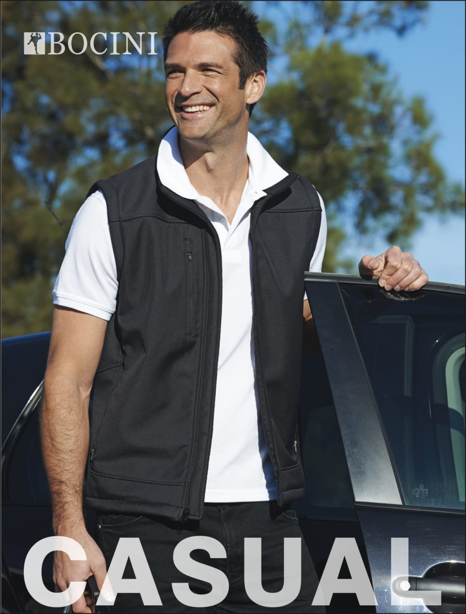 A black softshell vest on a smiling man, standing beside a car, featuring a logo.