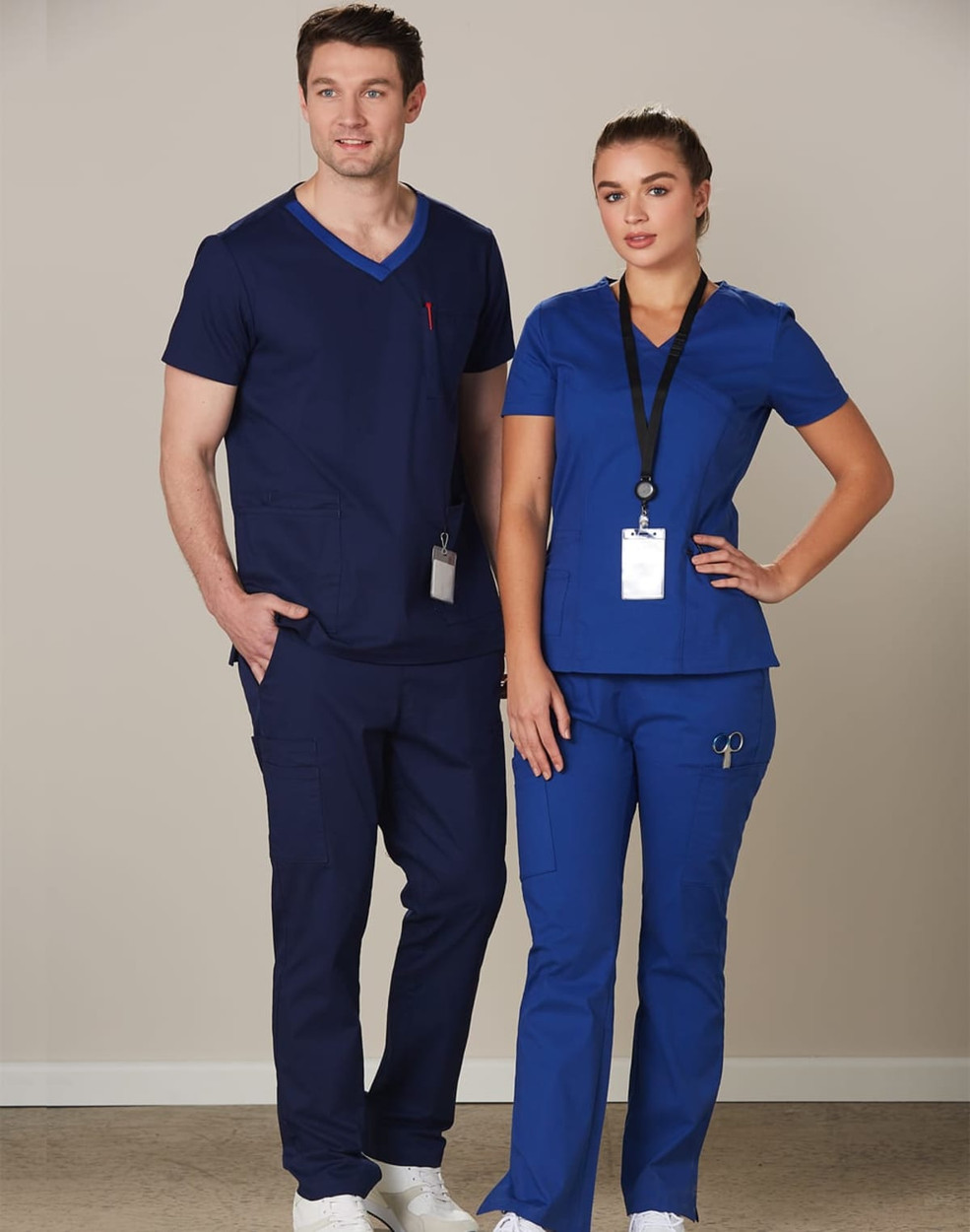 A man and woman wear solid colour scrub tops and pants in navy and blue, with badges and a logo present.