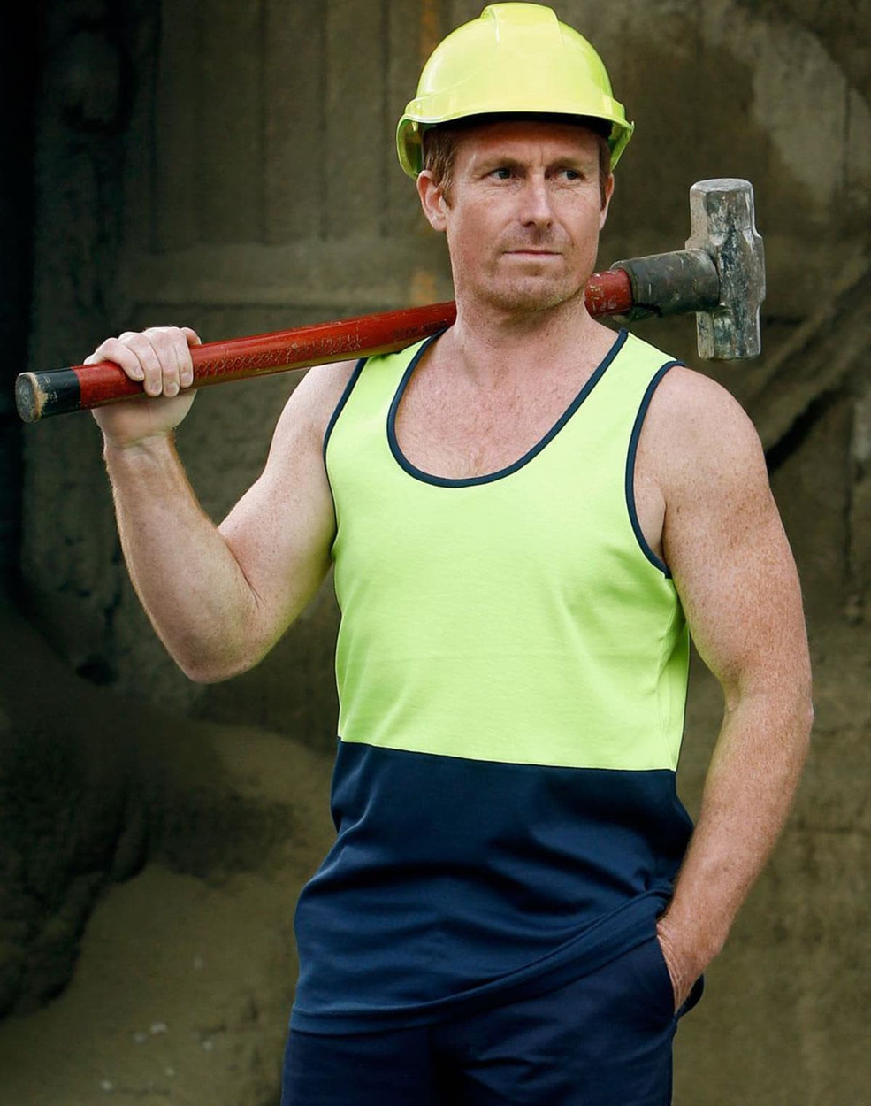 A man wearing a hi-vis yellow and navy safety singlet, holding a hammer over his shoulder, with a hard hat on.