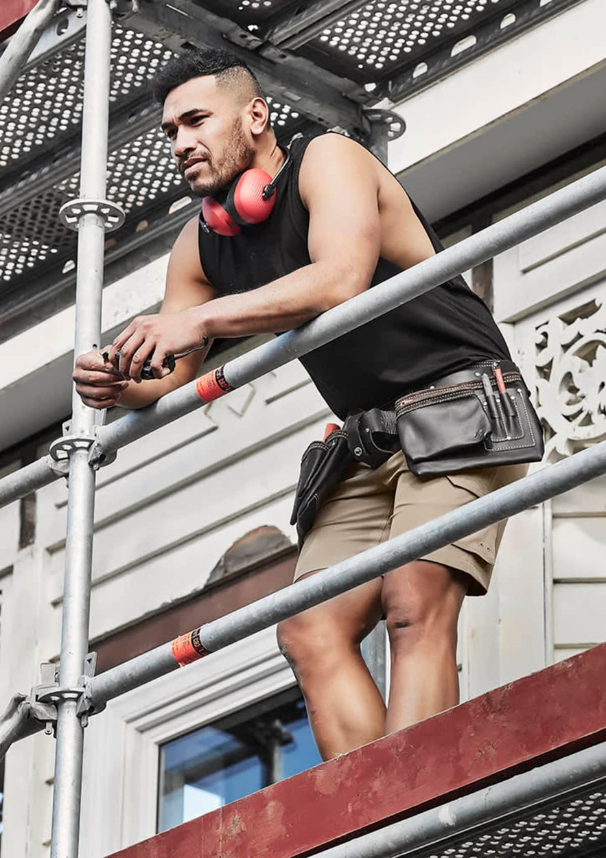 A man in a black sleeveless tee and shorts stands on scaffolding, wearing headphones and a tool belt.