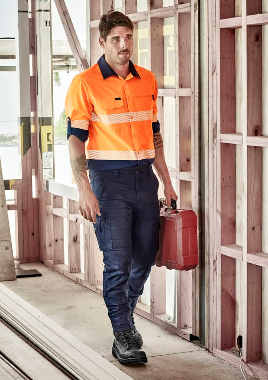 A man wearing an orange long sleeve segmented tape work shirt and navy pants, holding a red toolbox in a construction setting.