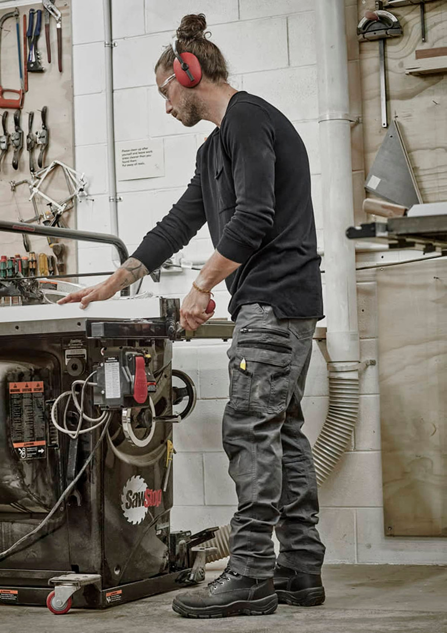 A man wearing black work pants stands by a workshop table, handling tools in a well-equipped space.