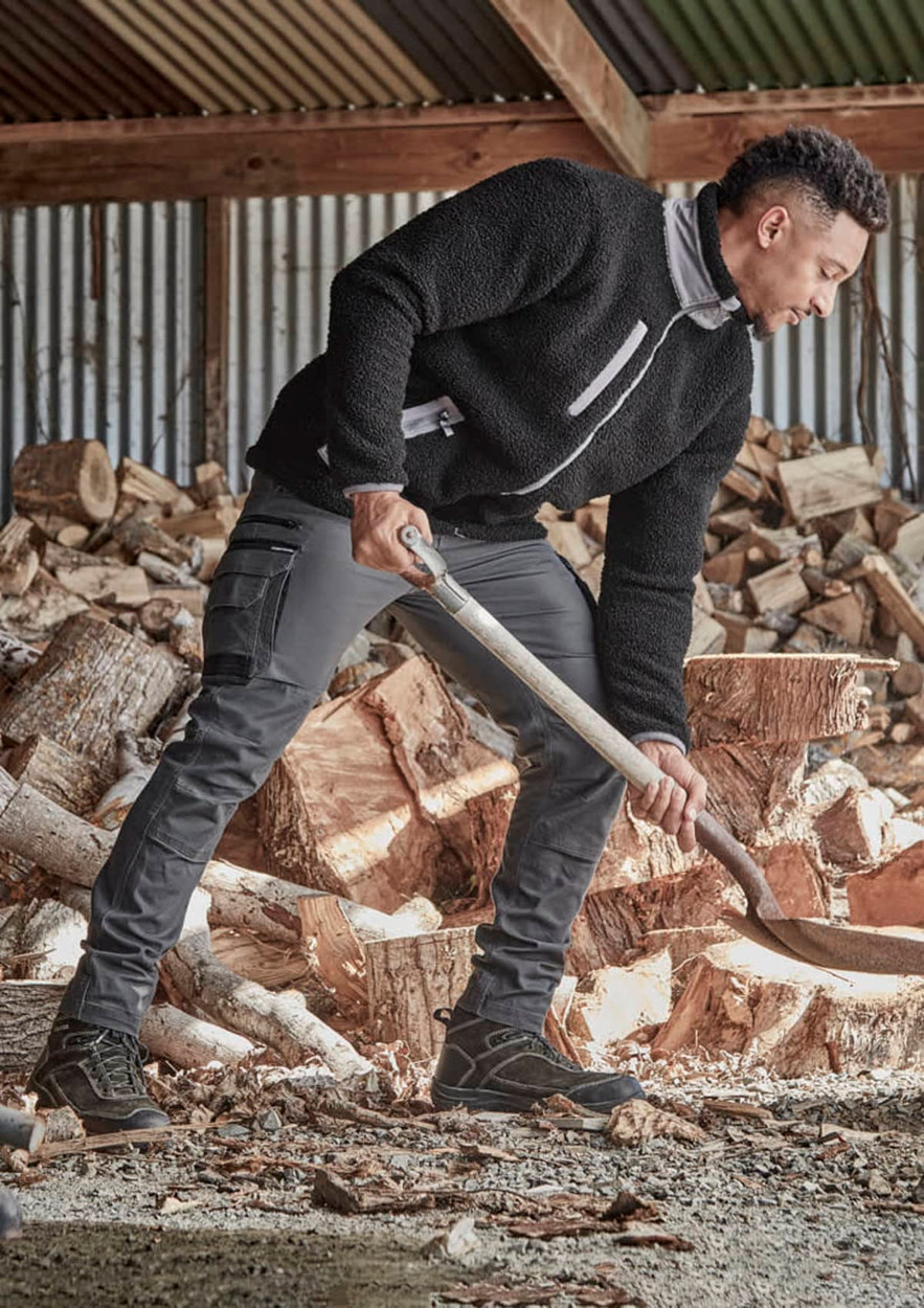 A model is wearing black work pants and a black fleece top while chopping wood in a rustic setting.