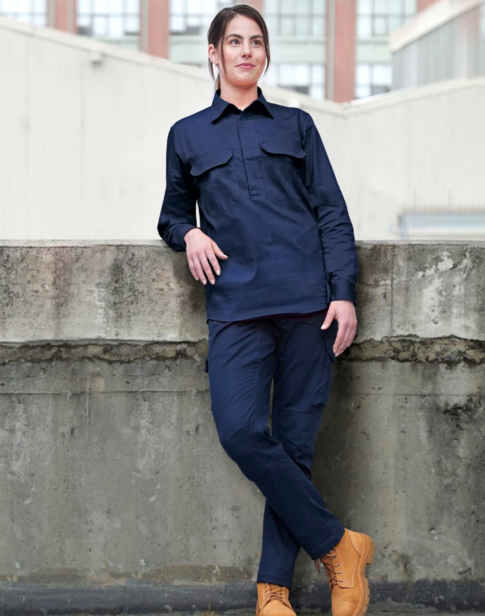 A navy long-sleeve work shirt with a closed front and two chest pockets, worn by a model. The model is leaning against a wall.
