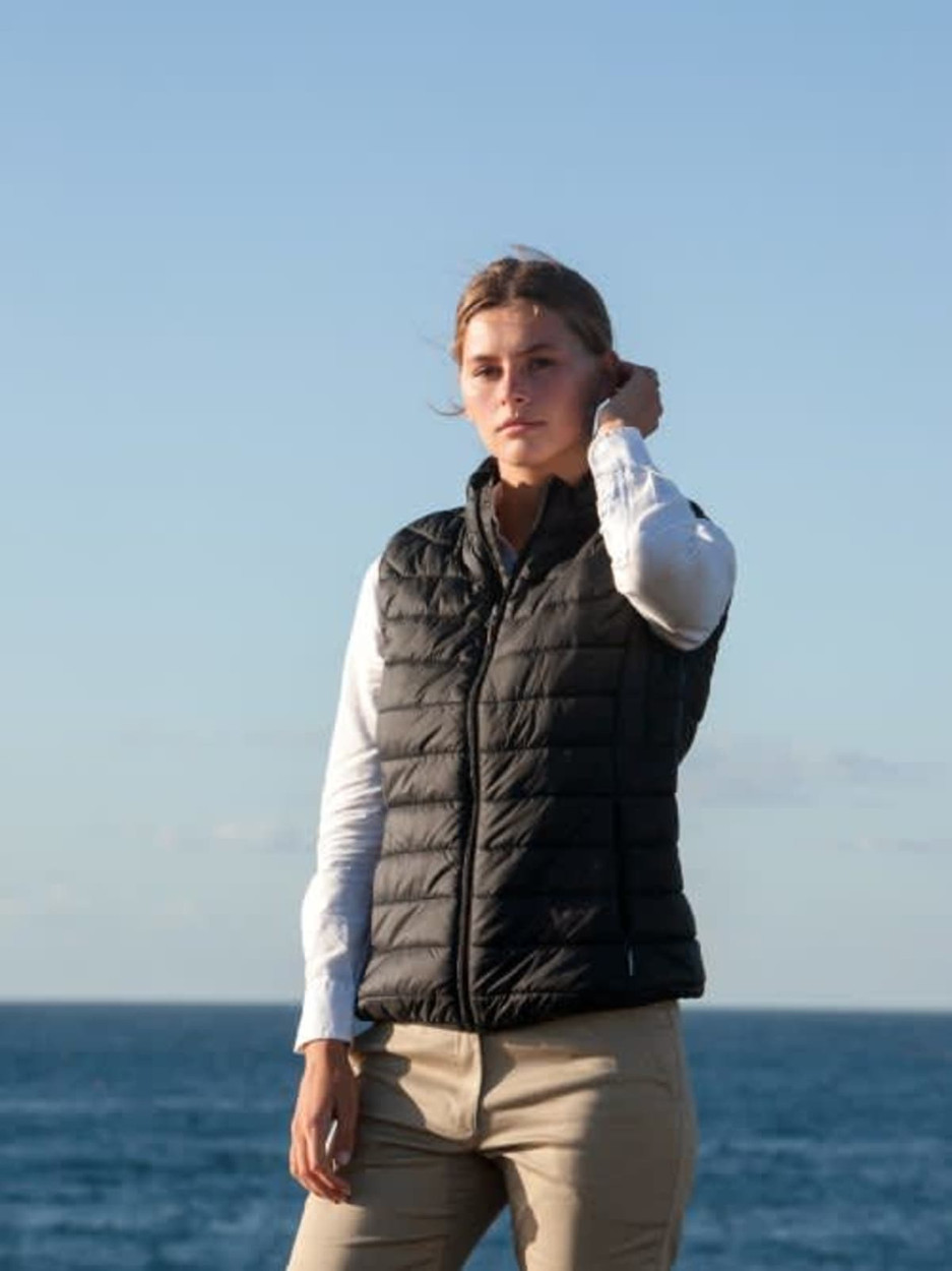 A woman stands outdoors wearing a black puffer vest over a white shirt, with the ocean in the background.