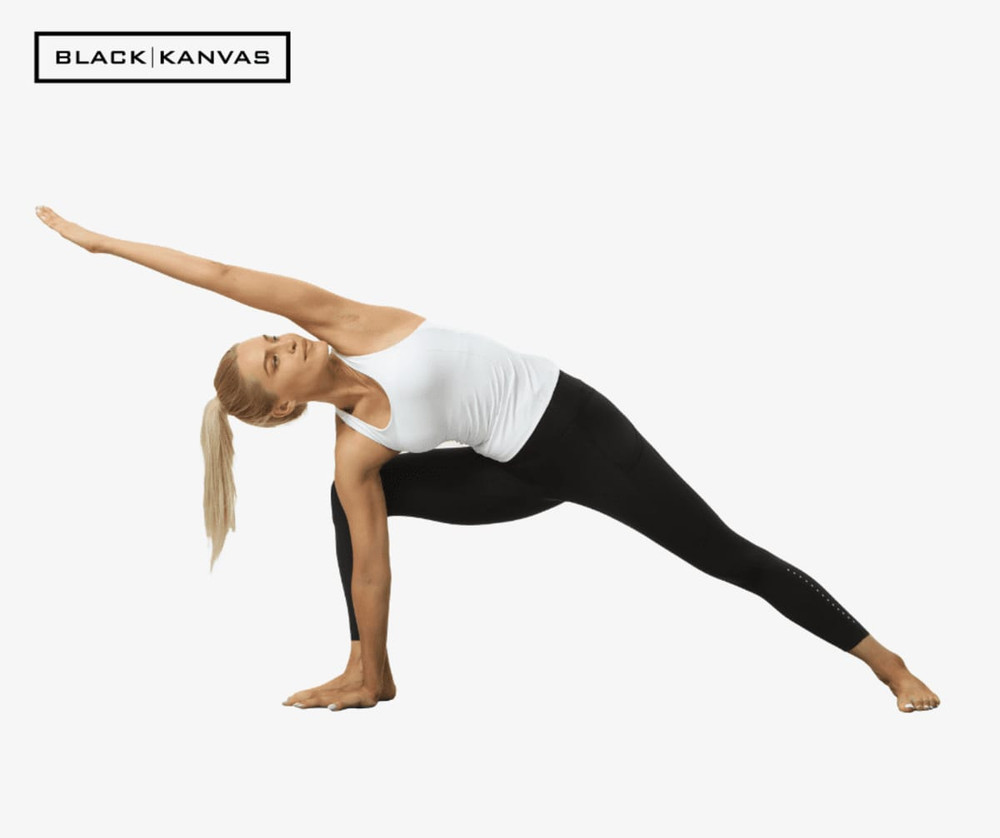 A person in a white top and black leggings performs a yoga pose against a plain background. The leggings feature a logo.