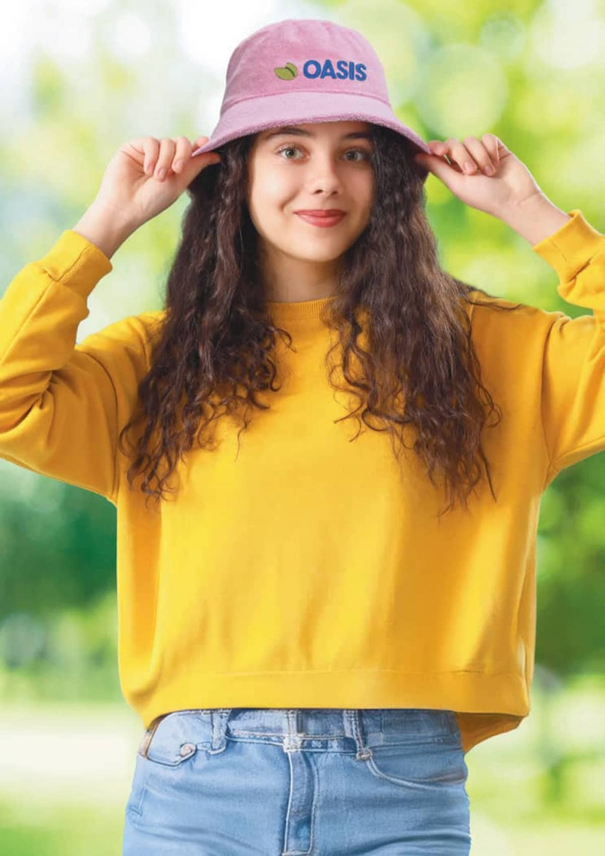 A pink terry towelling bucket hat with a logo, worn by a smiling person in a yellow sweatshirt against a green background.