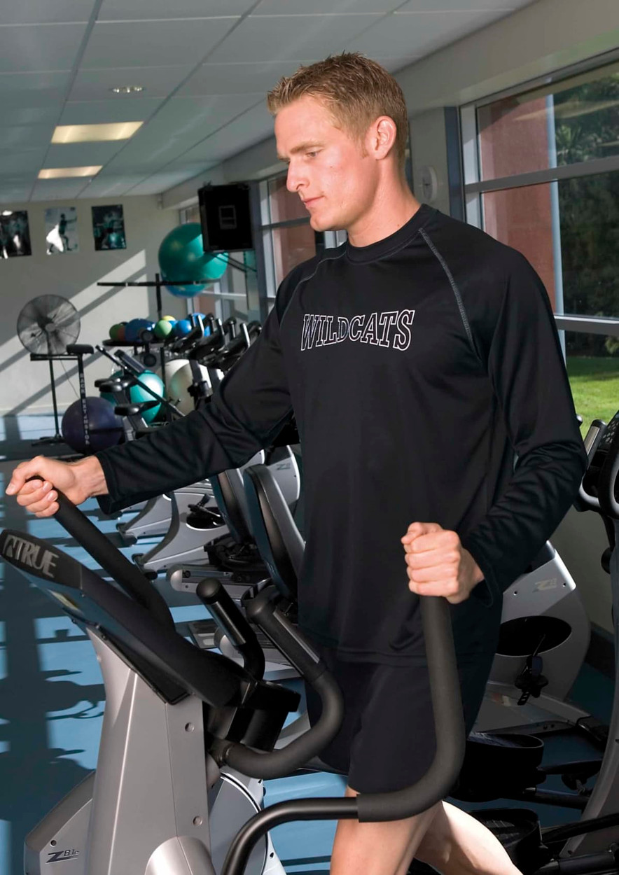 A man wearing a black long sleeve tee exercises on an elliptical machine in a gym setting. The shirt has a logo.