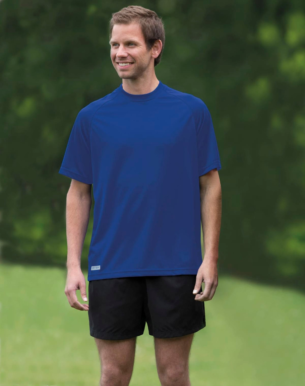 A man stands outdoors, wearing a blue Dri Gear Plain Raglan Tee and black shorts. The shirt has a logo on it.