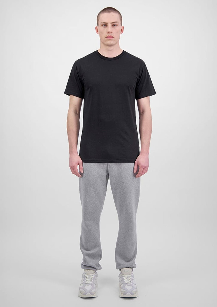 A person wears a black t-shirt and grey track pants, standing against a plain background. The outfit features branding.