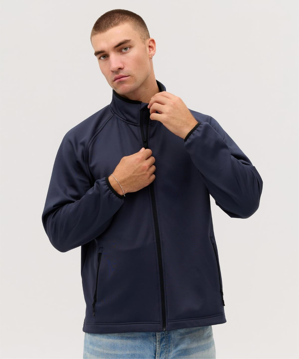 A man wearing a navy soft shell jacket, standing with his hands on the collar, featuring a logo.