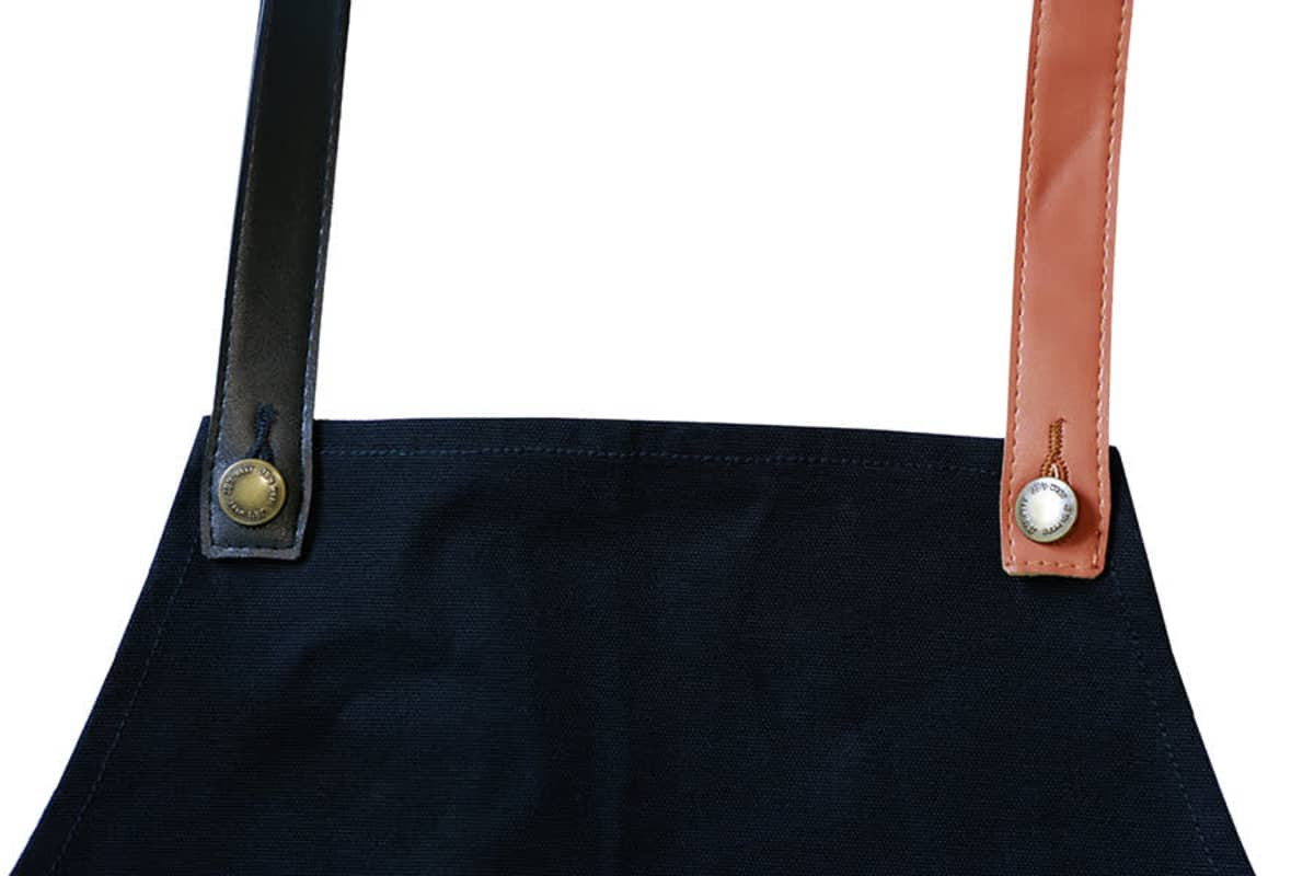 A black apron with changeable PU cross-back straps in black and brown, featuring metal buttons.